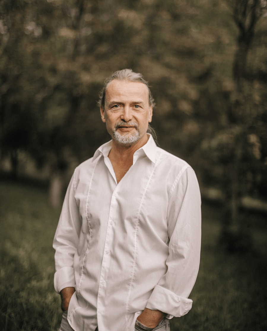 Reijk Nilson, Life Coach und Heiler aufgenommen in der Natur mit weissem Hemd die Hände in den Taschen, schaut in die Kamera und lächelt.
